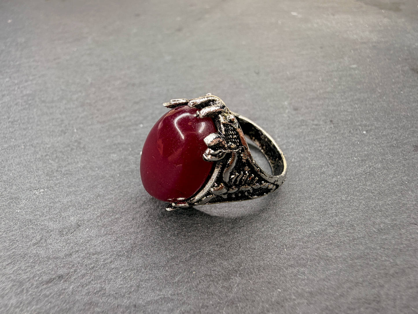 A silver-toned ring featuring a large, smooth, oval-shaped deep red cabochon gemstone, photographed on a textured gray surface.