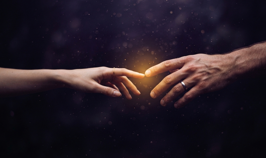 Two hands reaching toward each other in the dark, almost touching with a warm golden light between the fingertips