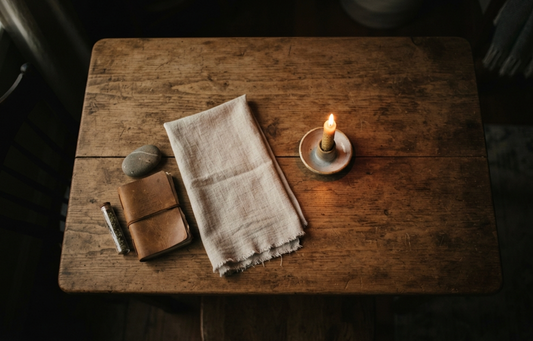 A rustic wooden table set with a leather journal, stone, herb vial, fringed linen, and glowing beeswax candle. The warm, dim lighting creates a calm atmosphere, visually representing the tools used to expect and prepare for a psychic reading