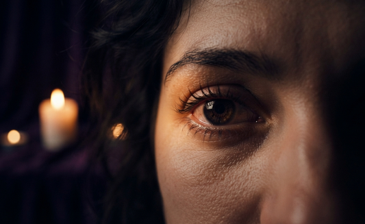 Close-up of a woman's right eye illuminated by a warm candle glow, featuring deep violet shadows and a mysterious mood.