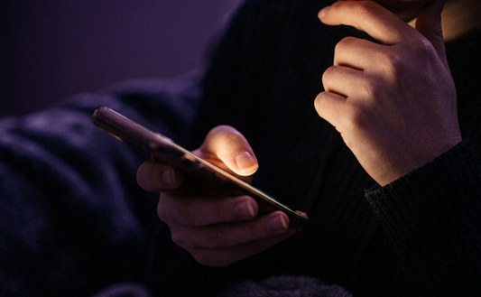 Close-up of a person holding a glowing phone screen in a dark room with deep violet shadows, representing late-night texting and wondering why an ex unblocked them.