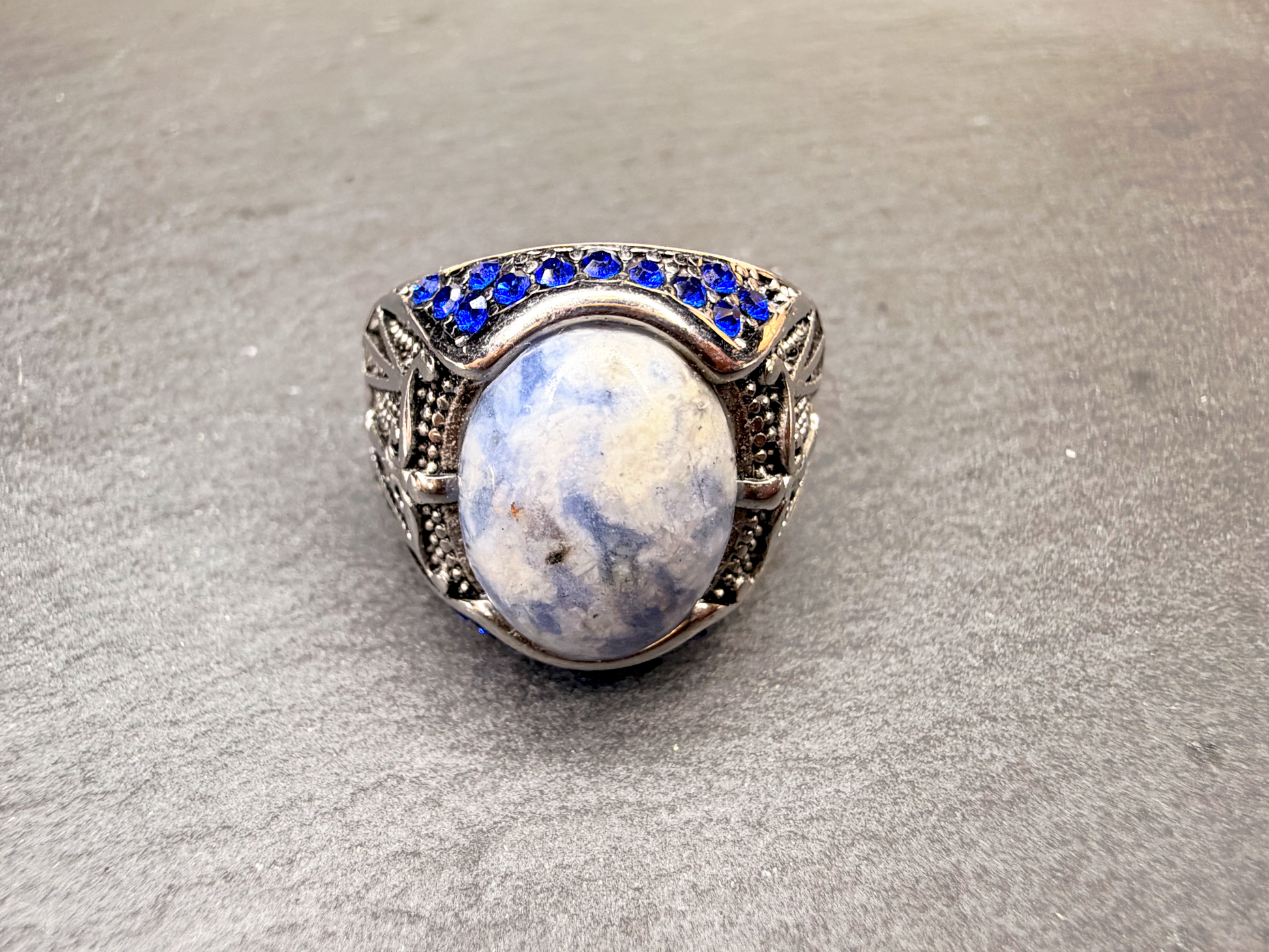 A silver-toned ring featuring a large oval cabochon stone with blue and white marbled patterns, accented by a row of small round blue stones along the top, set against a textured gray background.