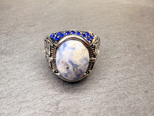 A silver-toned ring featuring a large oval cabochon stone with blue and white marbled patterns, accented by a row of small round blue stones along the top, set against a textured gray background.