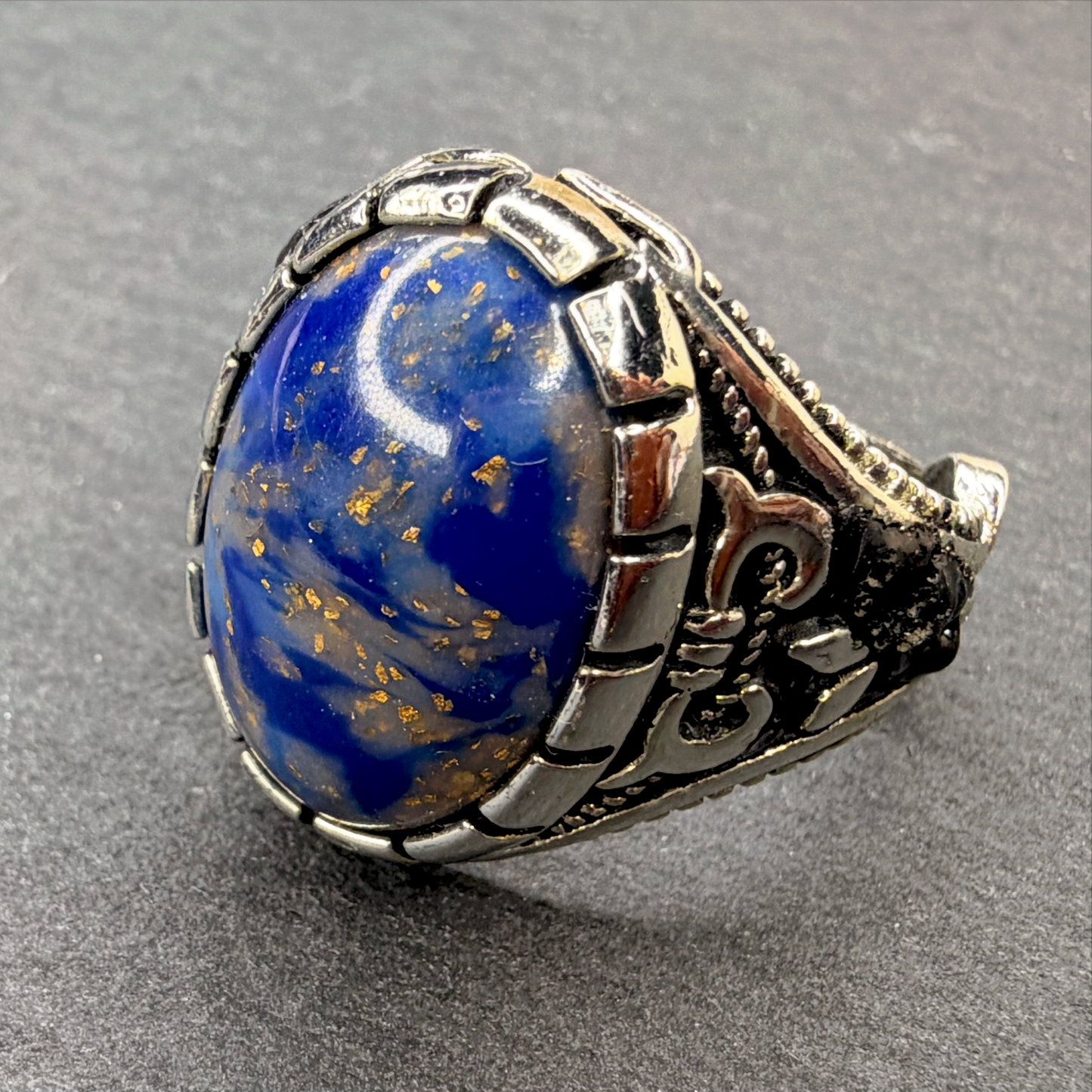 A silver-toned ring featuring a large oval cabochon lapis lazuli stone with visible golden pyrite flecks, set in an ornate textured silver band on a dark gray background.