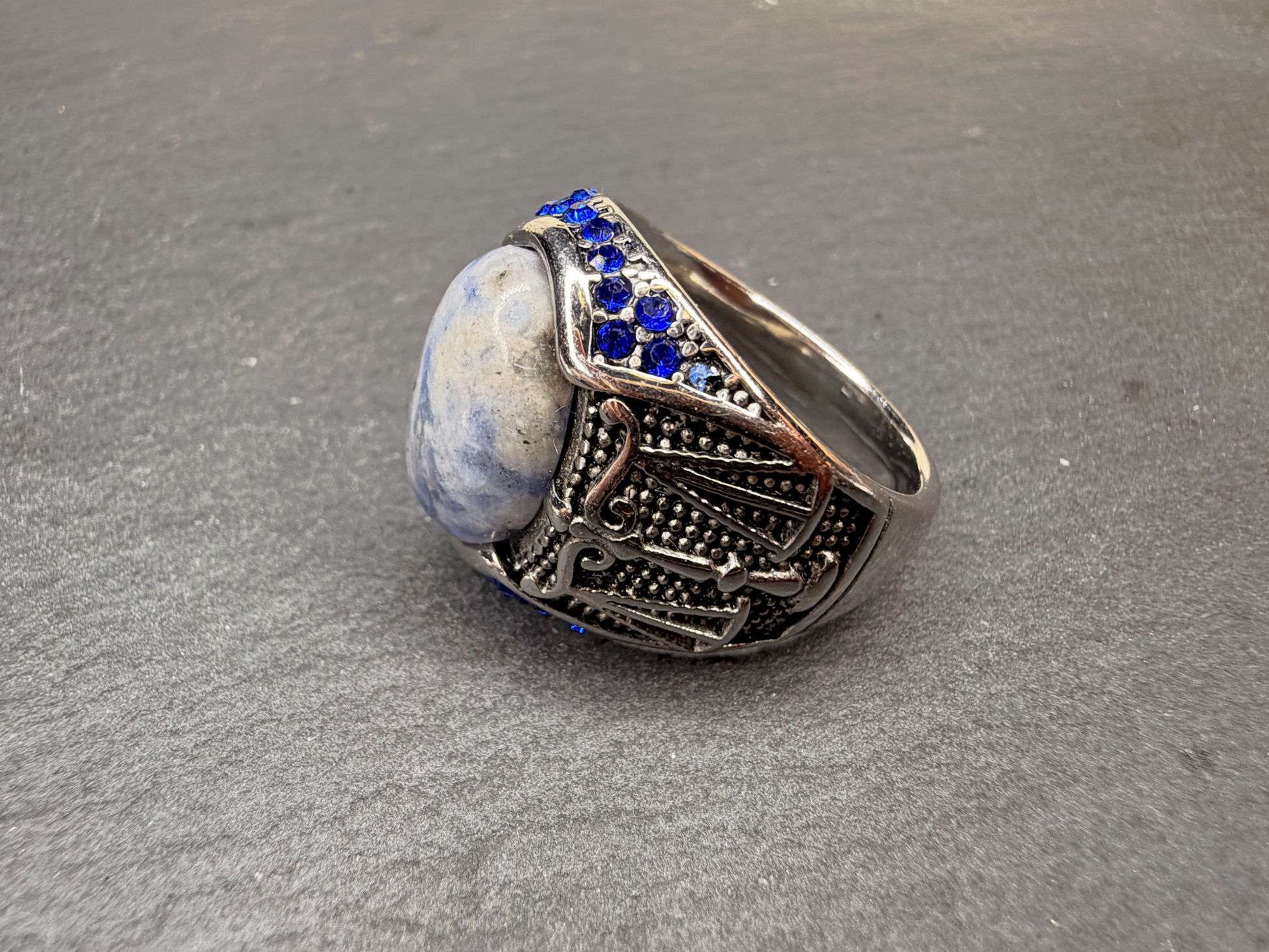 A silver-toned signet ring featuring a large oval blue and white stone, accented with small round blue stones along the top, set against a textured gray background.