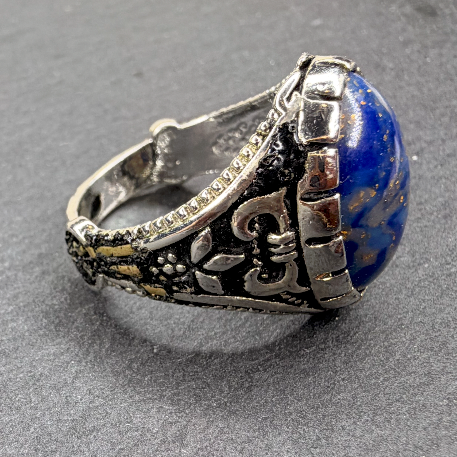 A silver-toned textured ring featuring a large, polished blue lapis lazuli stone, photographed on a dark gray textured background.