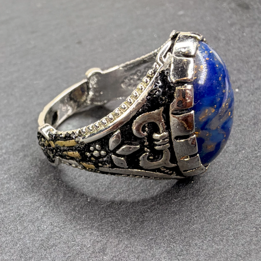 A silver-toned textured ring featuring a large, polished blue lapis lazuli stone, photographed on a dark gray textured background.