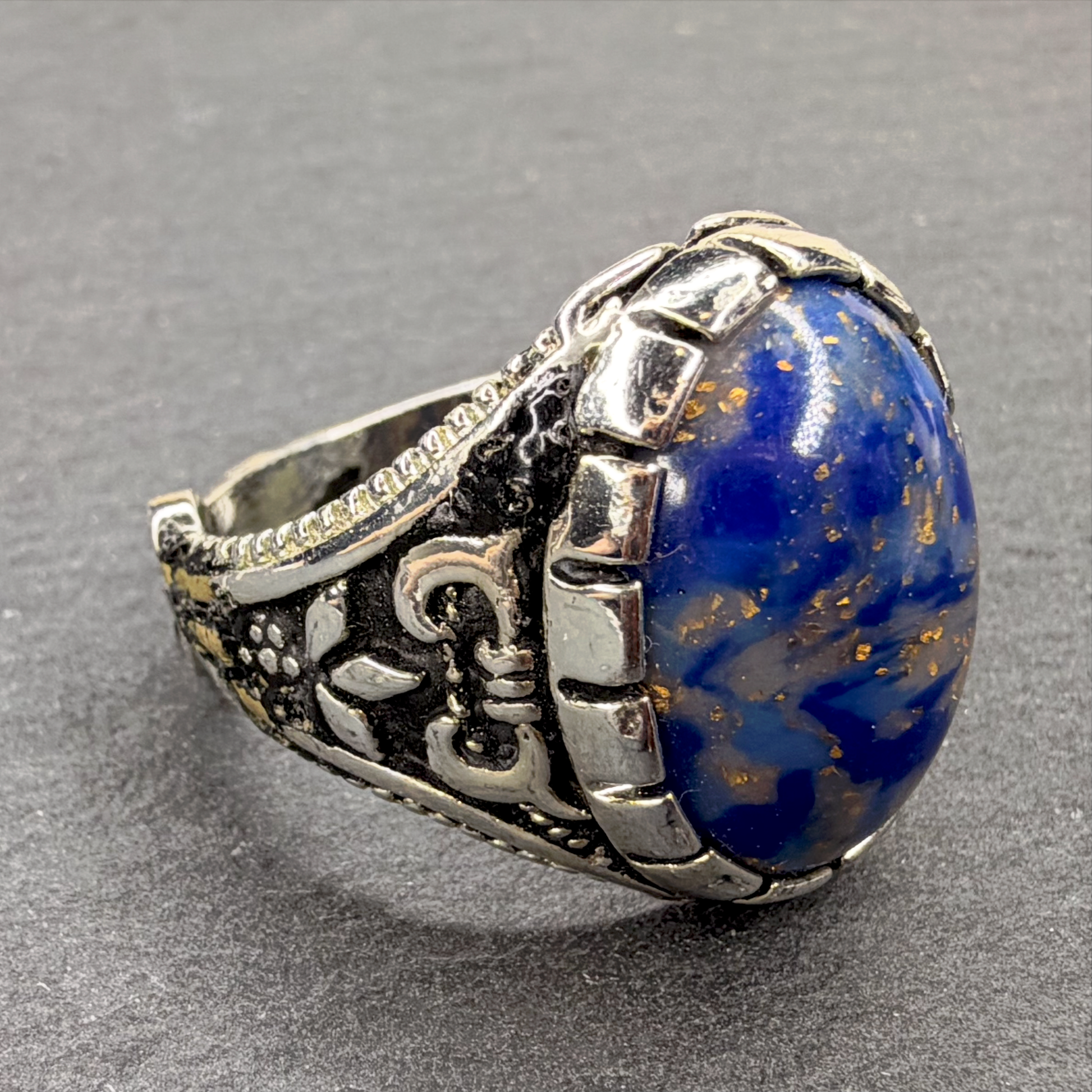 A silver-toned engraved ring featuring a large oval cabochon stone with swirling blue, white, and gold flecks, set against a neutral gray background.