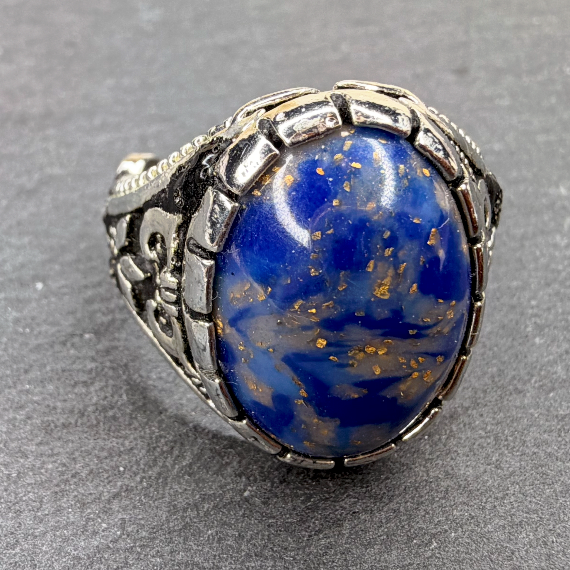 A silver ring featuring a large oval cabochon of blue lapis lazuli with visible gold pyrite flecks, set in an ornate silver band on a textured gray background.