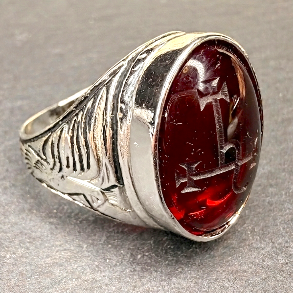 A silver signet ring with an oval dark red cabochon stone featuring a carved cross design, resting on a textured gray surface.