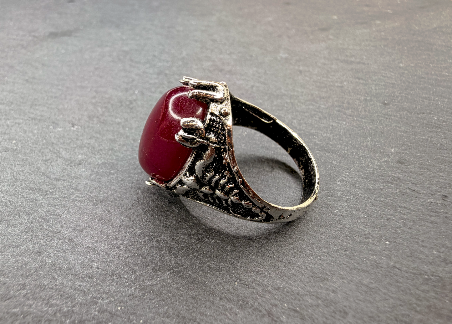A silver-toned ring with a textured, claw-like setting holding a smooth, oval red gemstone, photographed on a dark gray textured surface.