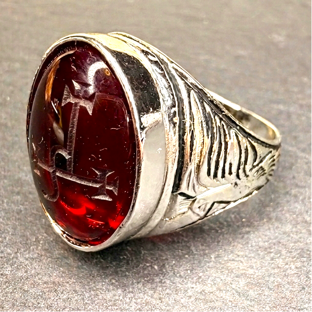 A silver-toned ring with a large oval dark red cabochon gemstone featuring a cross motif, resting on a textured gray surface.