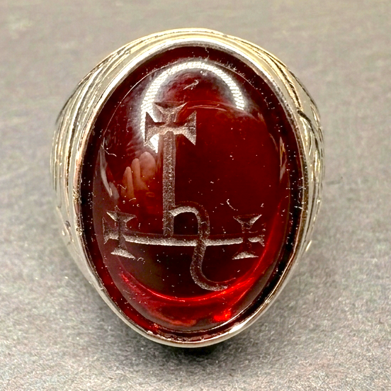 A silver-toned ring featuring an oval red cabochon stone with an engraved symbol, shown on a textured gray background.