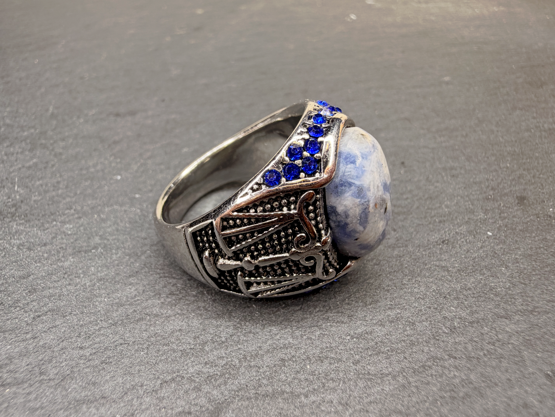 A silver-toned ring featuring a large oval blue and white stone, accented with small round blue stones along the top, set against a textured gray background.
