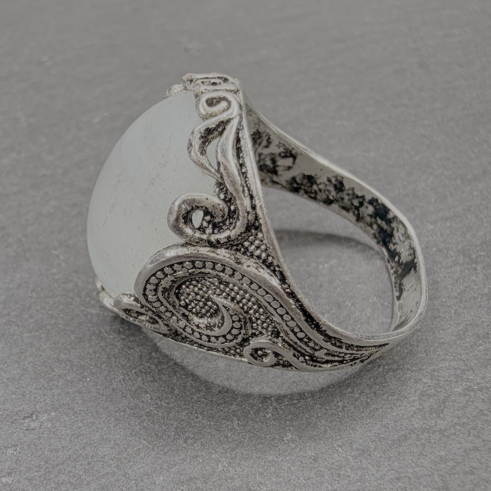 A silver-toned ring with an ornate paisley filigree design on a textured gray background.