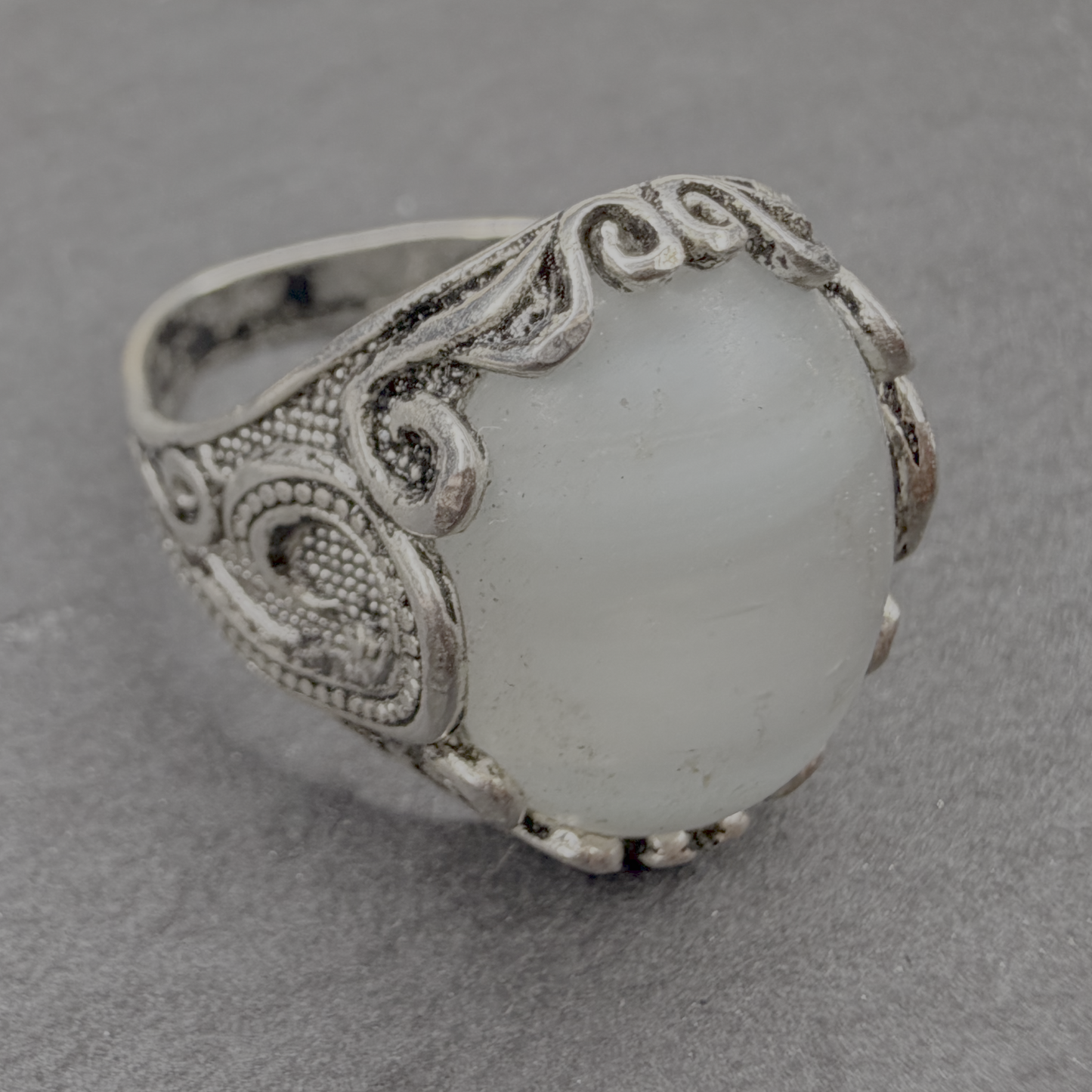 An ornate silver ring featuring a large, oval, milky white stone with subtle gray banding, set in a detailed filigree-style silver band, photographed on a textured gray surface.
