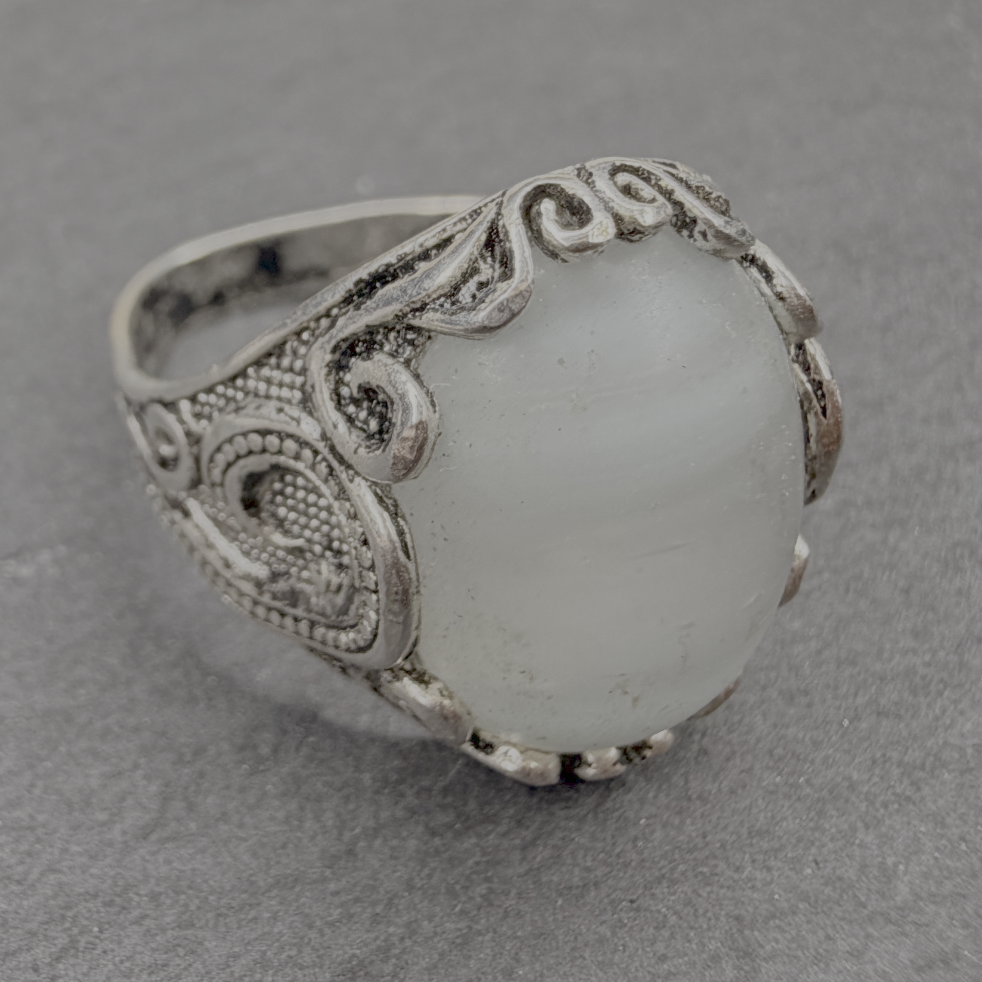 An ornate silver ring featuring a large, oval, milky white stone with subtle gray banding, set in a detailed filigree-style silver band, photographed on a textured gray surface.