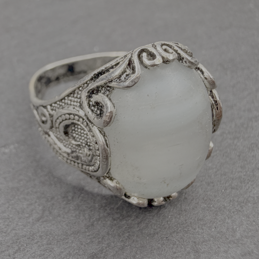 An ornate silver ring featuring a large, oval, milky white stone with subtle gray banding, set in a detailed filigree-style silver band, photographed on a textured gray surface.