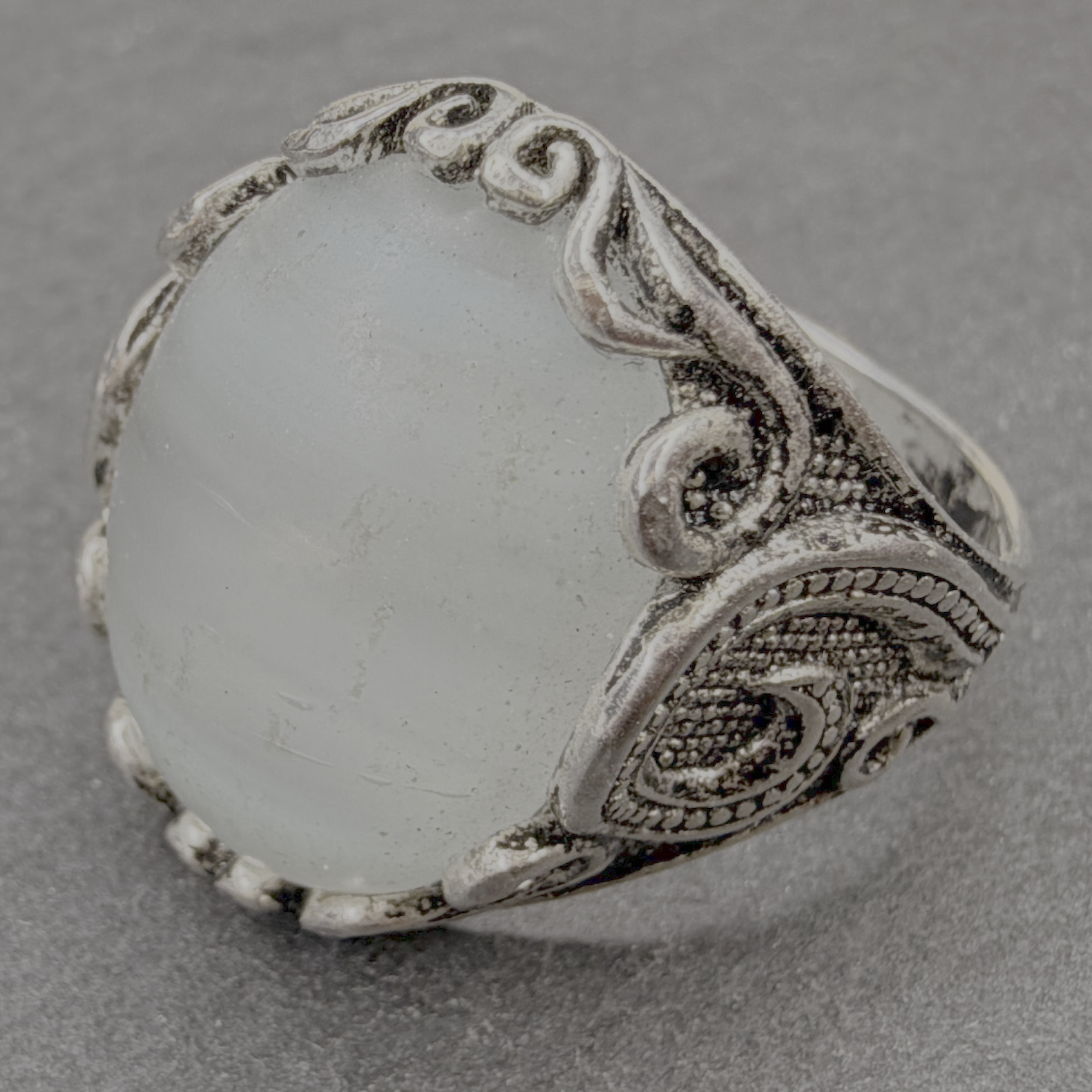 A silver ring with an oval milky white stone, featuring intricate filigree detailing on the band, set against a textured gray background.